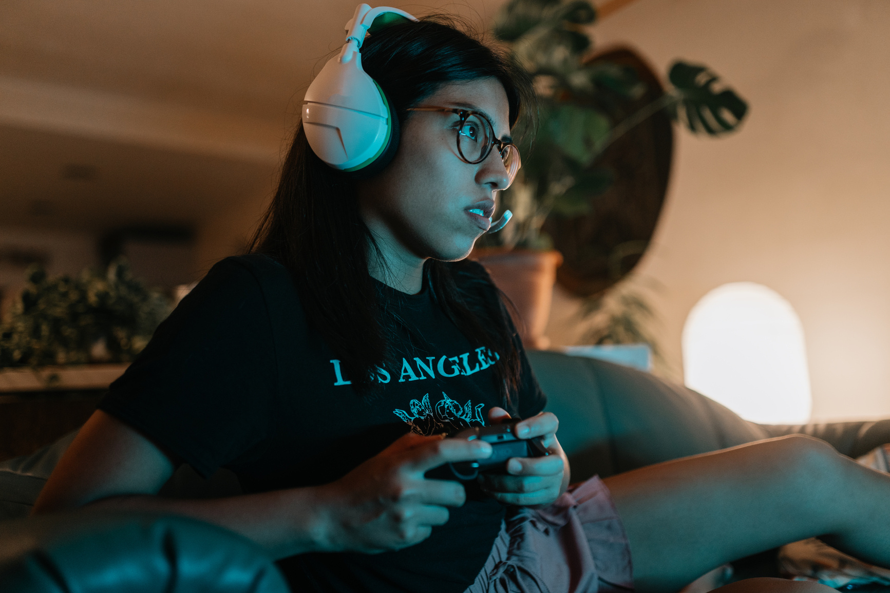 Una mujer con auriculares blancos sostiene un mando de videojuegos mientras mira la pantalla. Lleva anteojos, una camiseta negra y pantalones cortos de color rosa