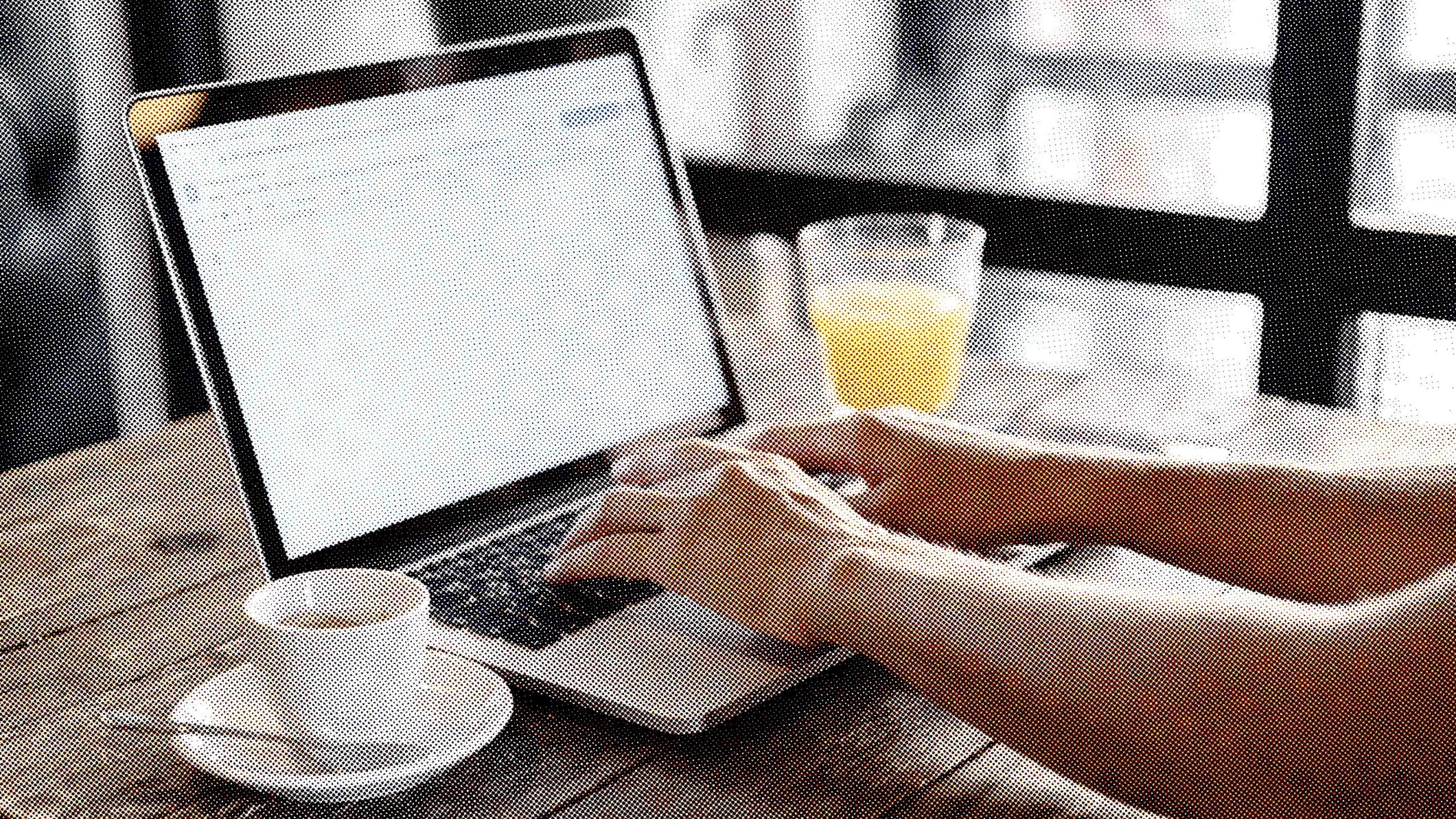 Un par de manos escribiendo en un equipo portátil, apoyado en una mesa de madera de una cafetería, junto a una taza de café y un vaso de jugo de naranja