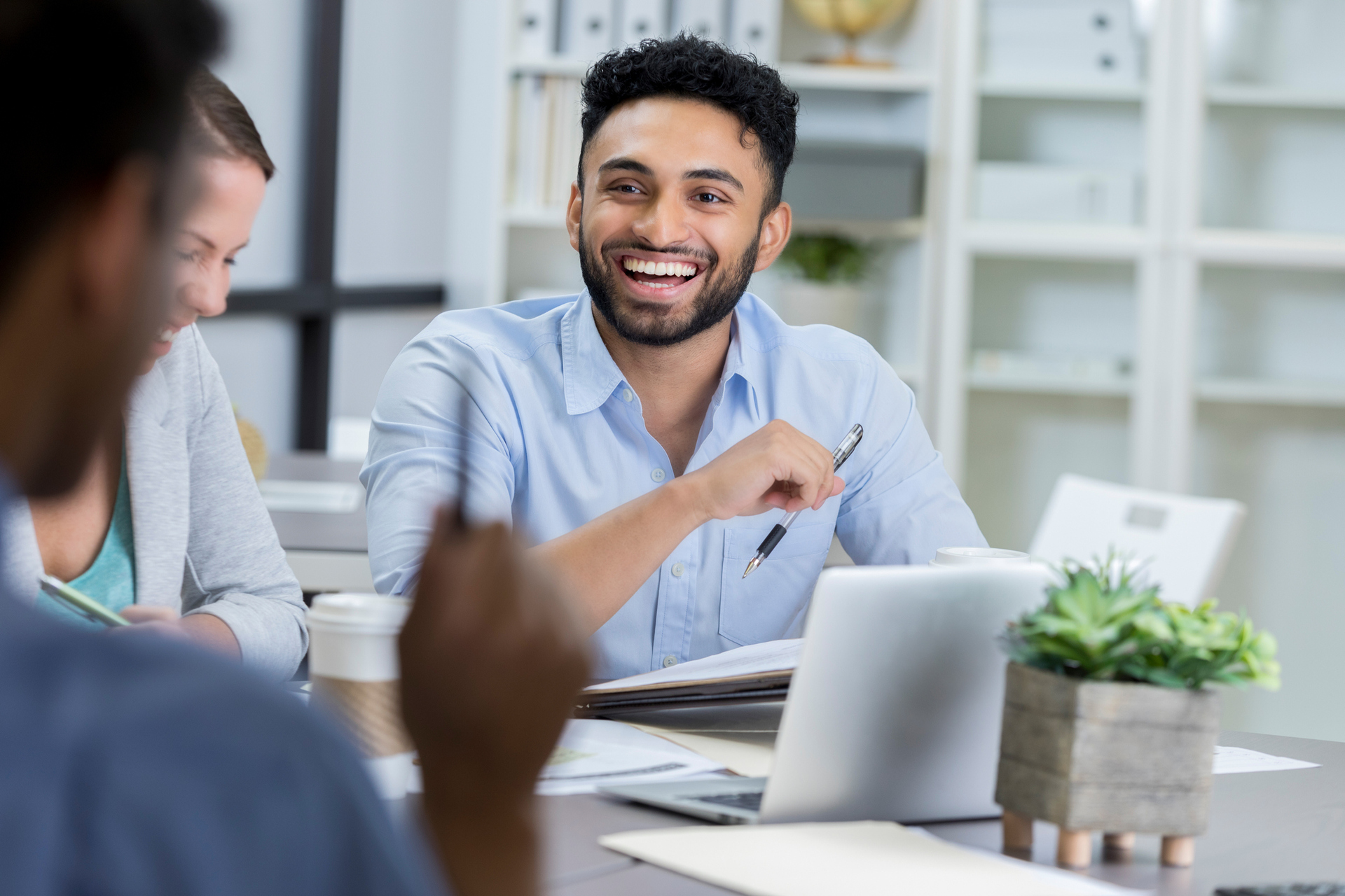 Homem usando camisa azul clara rindo junto a colegas dentro de um escritório